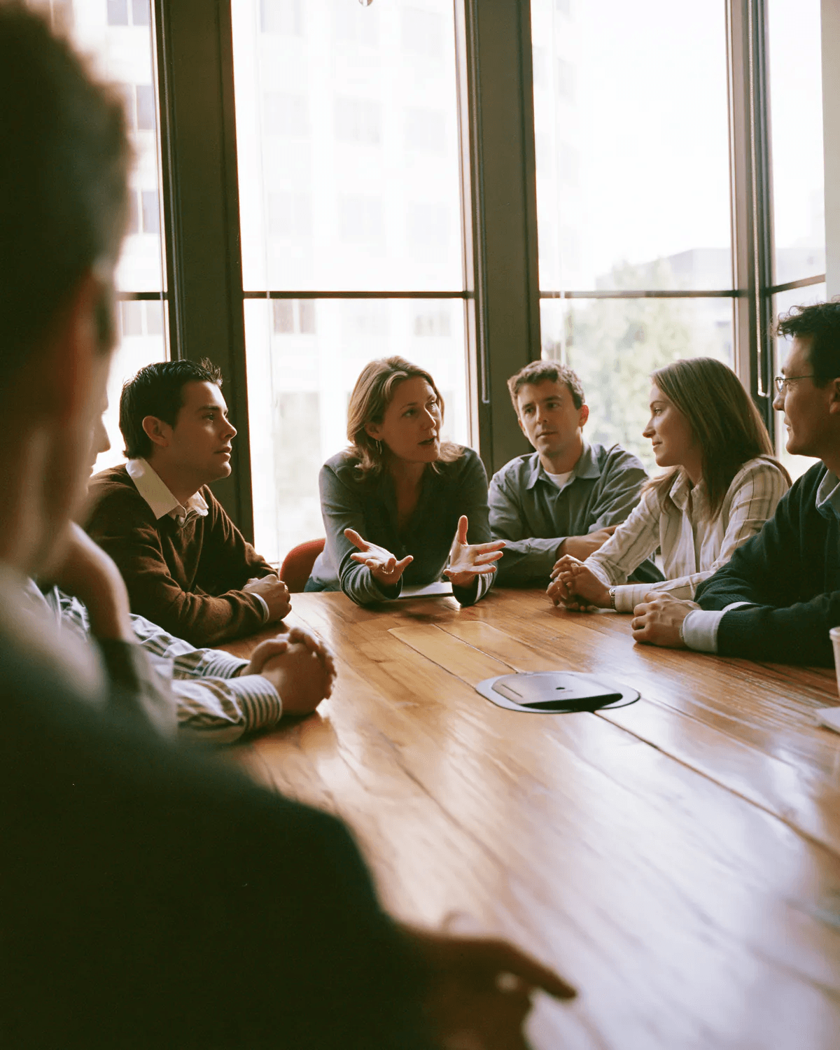A team in a meeting around a long wooden table, one person mid-sentence while the others listen